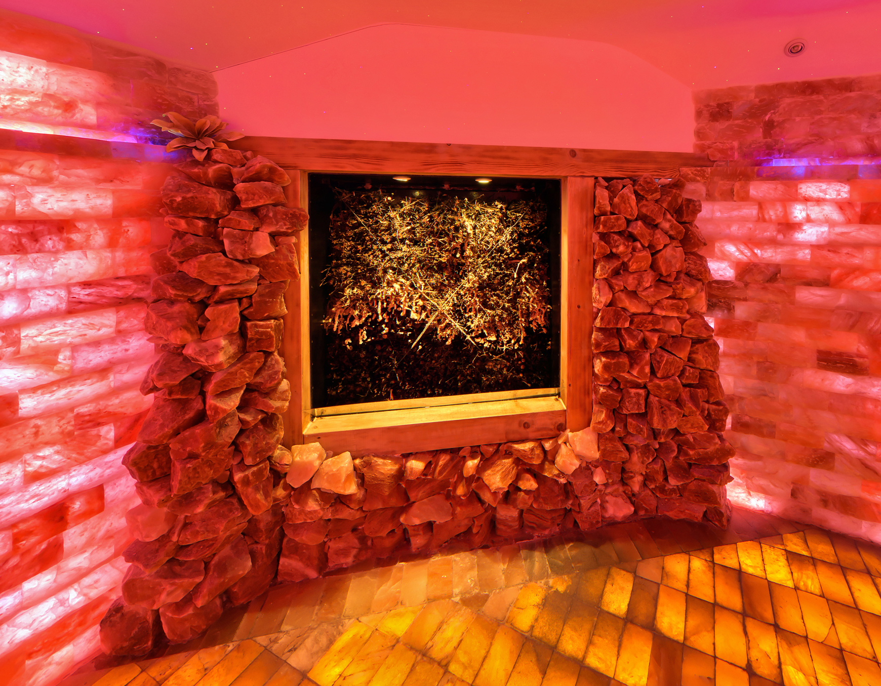 Raum mit warmem, rötlich-orangem Licht, dessen Wände aus Salzsteinen bestehen. In der Mitte befindet sich ein beleuchtetes Wandbild aus Kristallen oder Salzbrocken. Der Boden reflektiert das Licht und erzeugt eine gemütliche, entspannende Atmosphäre – typisch für eine Salzgrotte oder ein Salztherapie-Zimmer.