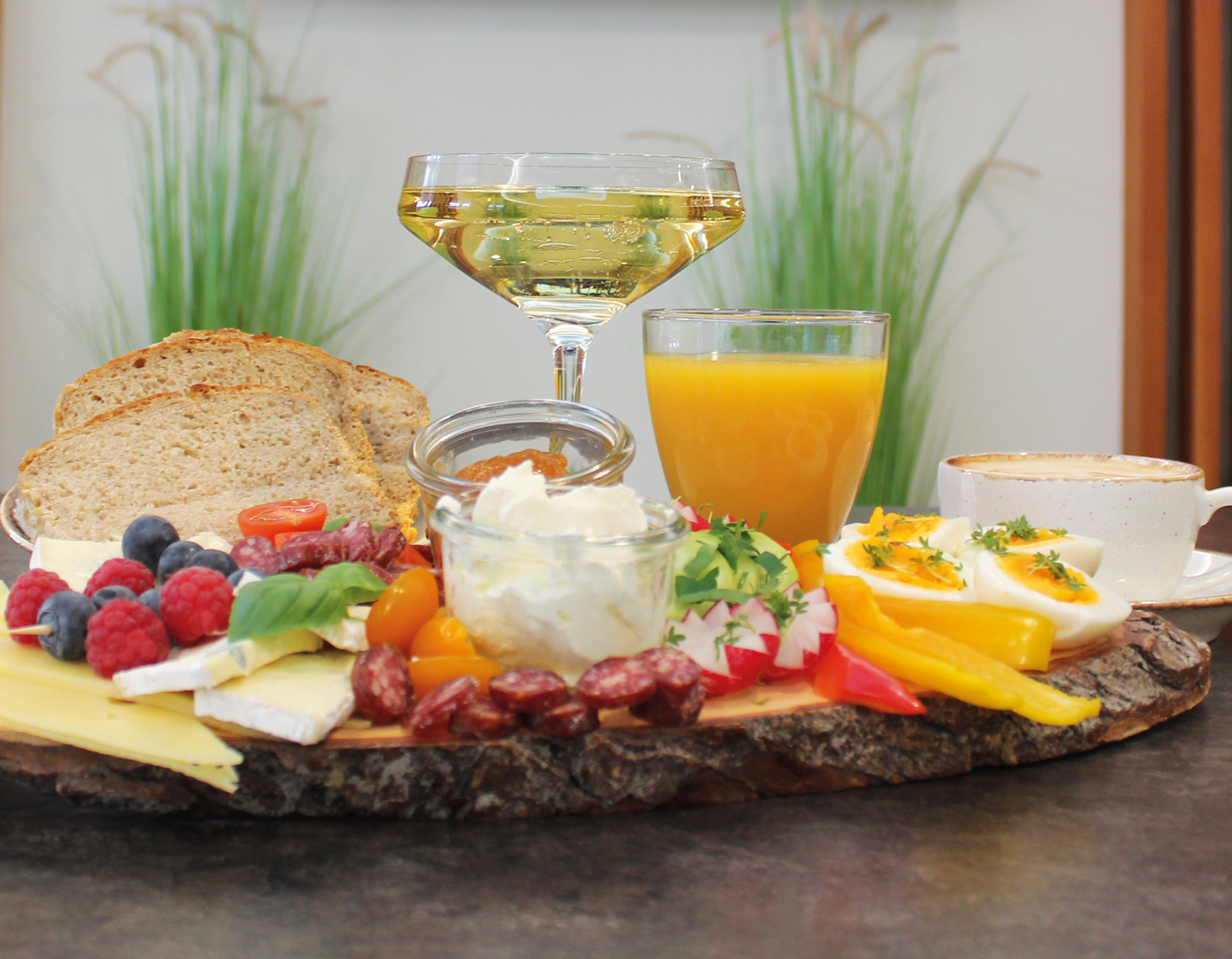 Eine rustikale Holzplatte mit verschiedenen Speisen, wie Gurke, gelbe und rote Paprika, Frischkäse und Marmelade im kleinen Einwegglas, Salami, Käse, gekochten halbierten Eier und Brotscheiben, dekorativ angerichtet. Im Hintergrund befindet sich ein Glas mit prickelnden Sekt, ein Glas Orangensaft und eine Tasse Kaffee.