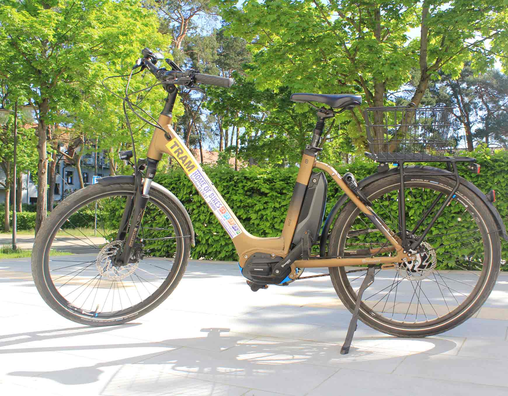 Fahrrad mit rotem Rahmen, schwarzem Lenker und Sitz, gelber Aufschrift HOLM, stehend im Ständer vor grüner Hecke und Bäumen.