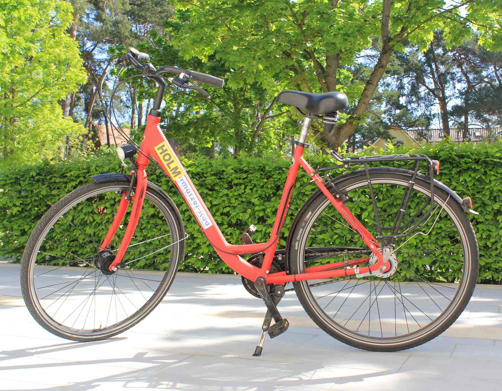 Fahrrad mit rotem Rahmen, schwarzem Lenker und Sitz, gelber Aufschrift ‚HOLM‘, stehend im Ständer vor grüner Hecke und Bäumen.