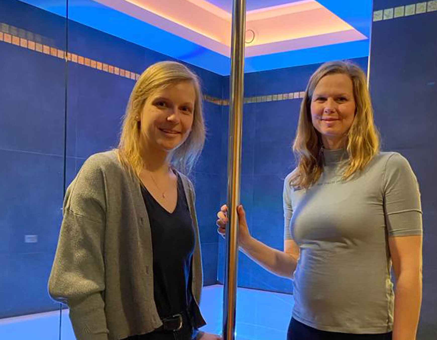 Zwei lächelnde Frauen stehen nebeneinander in einem modern beleuchteten Raum mit blauer Wand und LED-Lichtleisten.