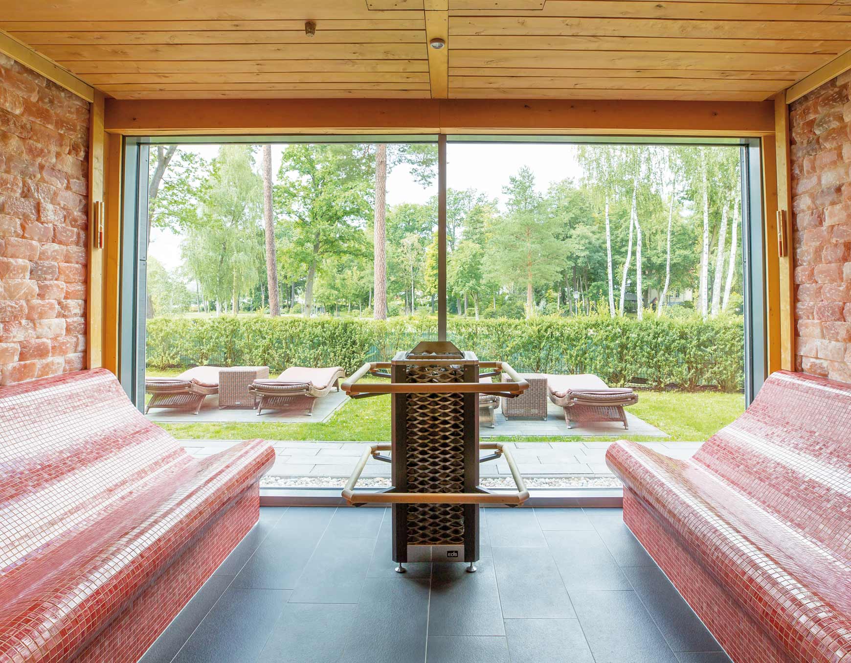 Aus  SalzSauna in der PanoramaSauna schaut man direkt auf den grünen Außenbereich mit braunen Liegen. Die Glasfront erhellt die Sauna mit natürlichem Sonnenlicht. Rechts und links von dem Metallfarbenen Saunaofen befinden sich jeweils rote, gewölbte Bänke aus kleinen Mosaikfliesen. Die Decke ist mit Holz abgedeckt und die Wände mit Salzsteinen versehen. 