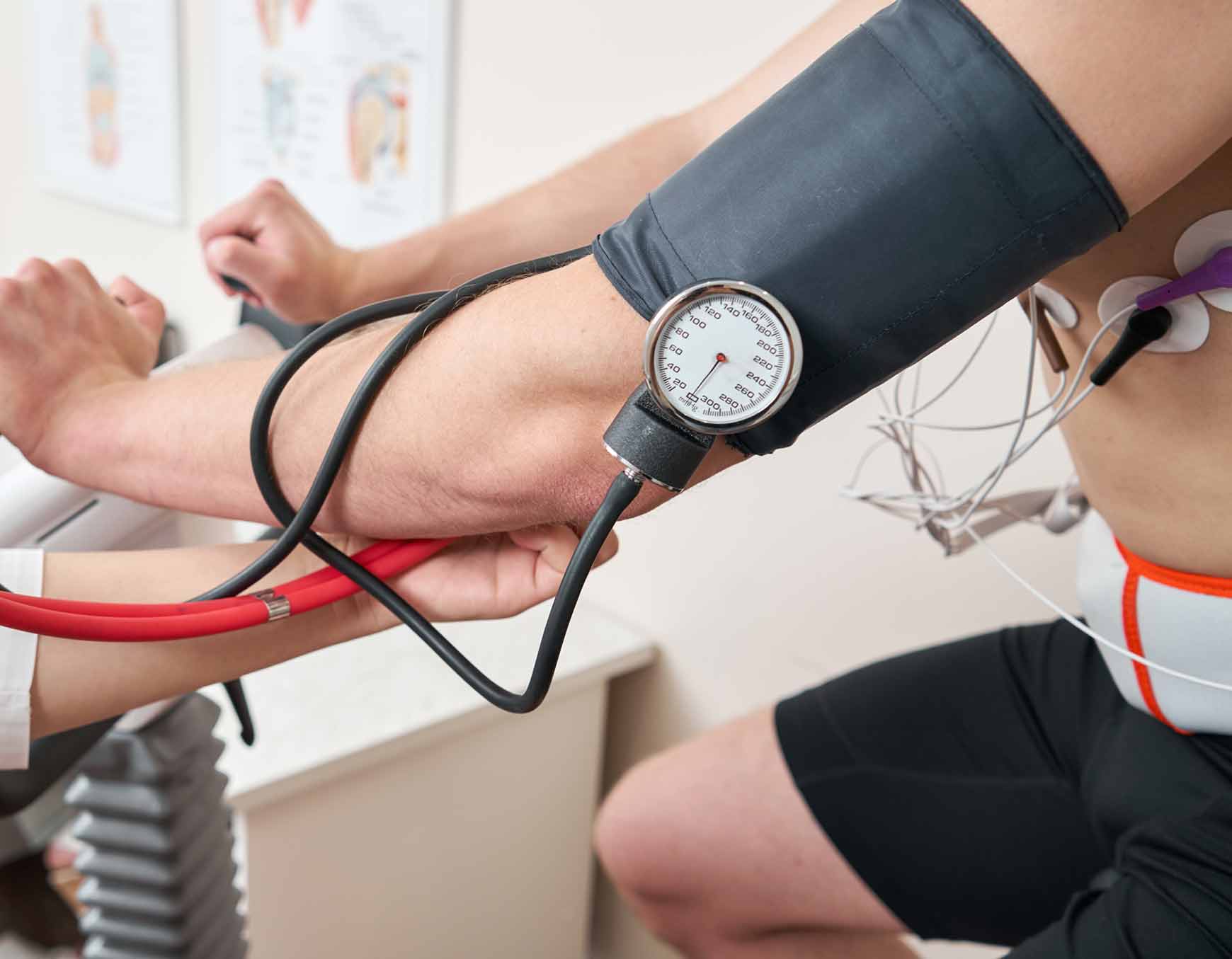 Ein Ausschnitt von einem Bild, wie ein Mann, Oberkörperfrei in einer schwarzen Sporthose auf einem Ergometer mit einem Blutdruckmessgerät am linken Oberarm und EKG Messgeräte am Oberkörper.
