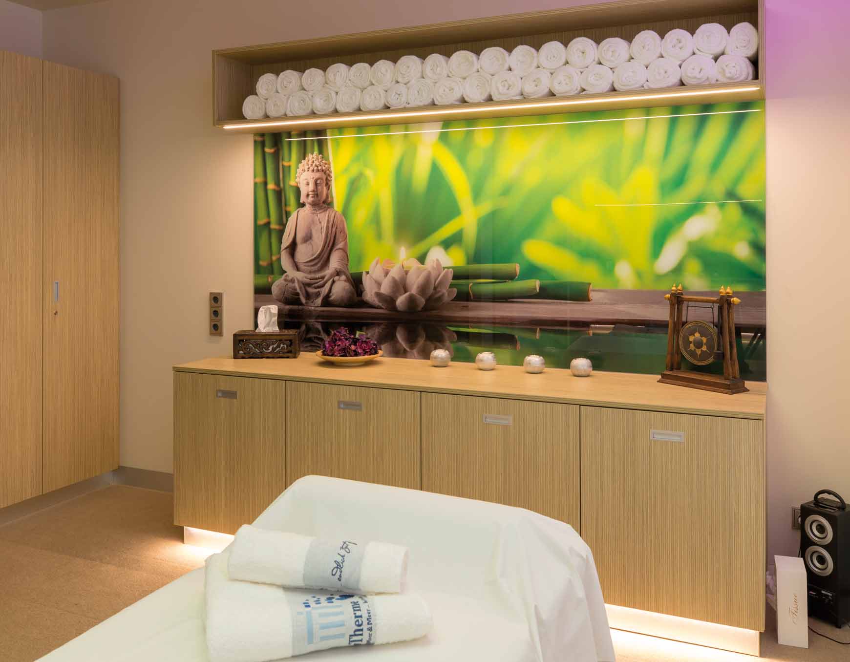 Ein hell erleuchteter Therapieraum mit Cremefarbenden Teppich. Im unteren Bereich des Bildes befindet sich eine Massageliege bedeckt mit einem weißen Laken und weißen Handtüchern mit der Aufschrift SaarowTherme. Im Hintergrund befindet sich eine helle Holzkommode mit Dekorativen Elementen wie einer Taschentücherbox, eine Schale mit Lila Blumen, 4 Teelichterständer und einem Chinesischen Gong Becken. Über der Kommode hängt ein hell grünes Bild mit einem Buddah, darüber ein Hängeschrank mit weißen Handtüchern