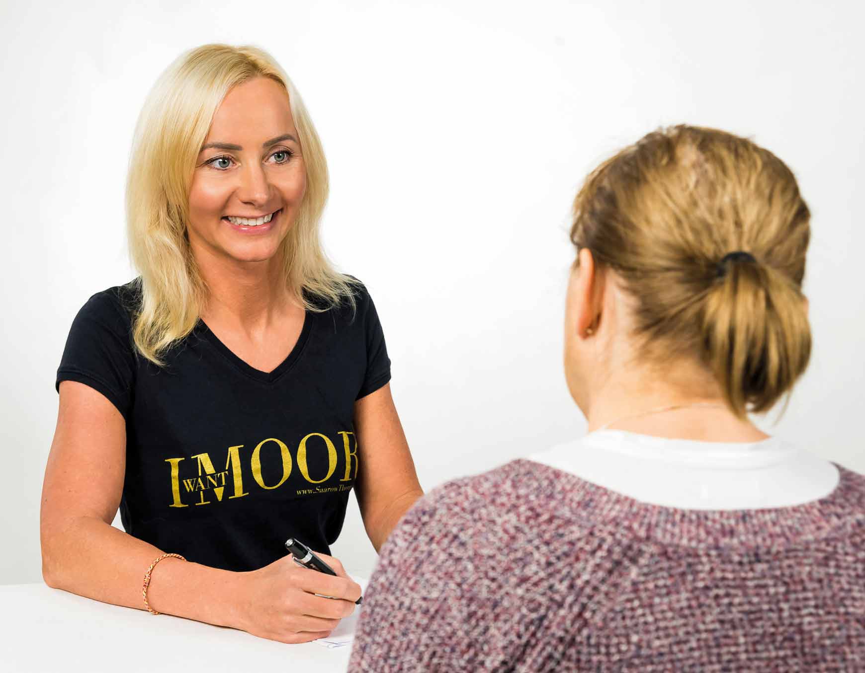 Abgebildet sind zwei Frauen in einem Beratungsgespräch. Die Patientin sitzt mit dem Rücken zur Kamera und trägt einen lilafarbenen Strickpullover. Das Gesicht der Therapeutin zeigt zur Kamera. Die Frau hat blonde Haare und ein schwarzes " I Want Moor" der SaarowTherme an. Sie lächelt die Patienten freundlich an. 