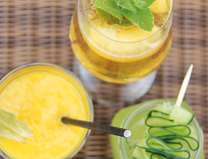 Drei farbenfrohe Sommergetränke in Gläsern – ein gelber Smoothie mit Physalis, ein goldener Drink mit Minze und Ananas sowie ein grüner Cocktail mit Gurkenspirale – auf einem geflochtenen Untergrund.