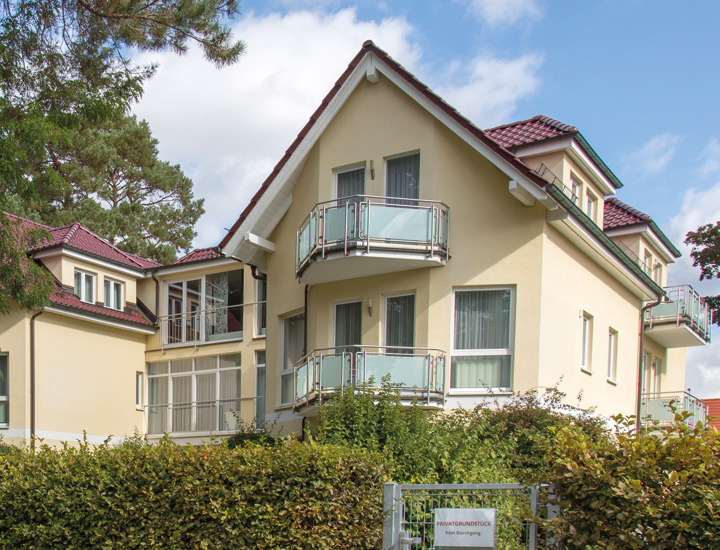 Zweistöckiges hellgelbes Wohnhaus mit roten Dächern und kleinen Balkonen, umgeben von Grünpflanzen und Bäumen unter blauem Himmel.