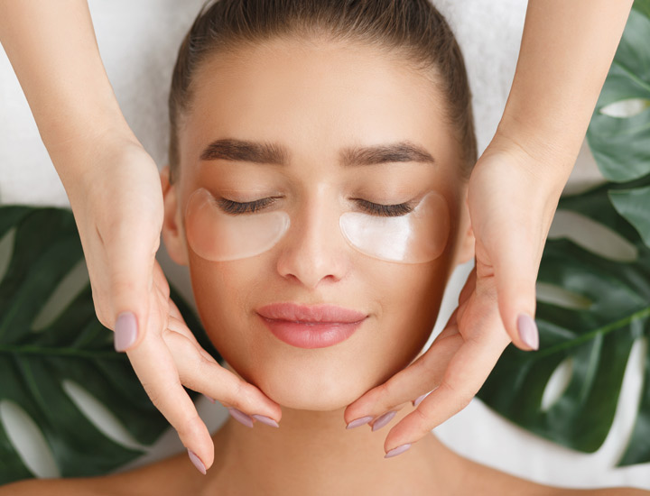 Eine junge Frau mit geschlossenen Augen liegt entspannt da, während transparente Augenpads unter ihren Augen haften. Sanfte Hände rahmen ihr Gesicht, das von gepflegten Augenbrauen und leichtem Make-up betont wird.