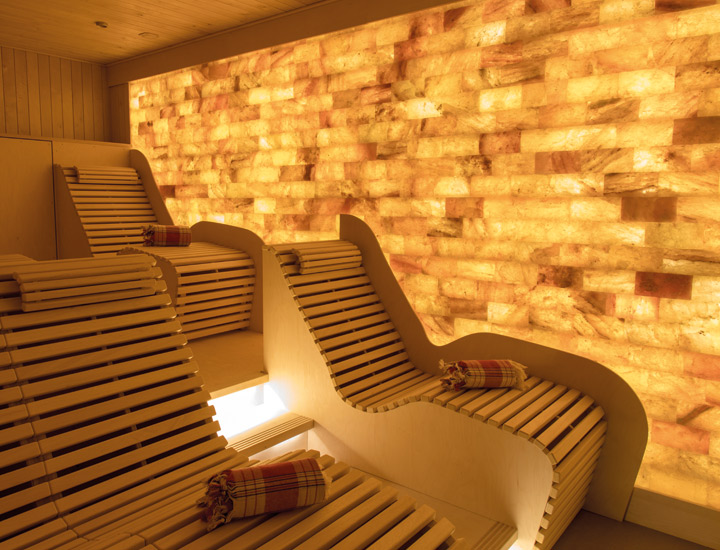 Sauna mit beleuchteter Salzsteinwand, ergonomisch geformten Holzliegen, die mit aufgerollten Hamamtüchern bestückt sind.
