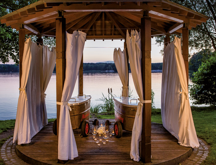Pavillon am See mit seitlichen Vorhängen drapiert für Privatsphäre. In der Mitte vom Pavillon stehen zwei Metallwannen auf Räder, die mit einem Deckel verschlossen sind. Zwischen den Wannen sind mehrere Kerzen auf den Boden verteilt worden, die für romantische Stimmung sorgen. 