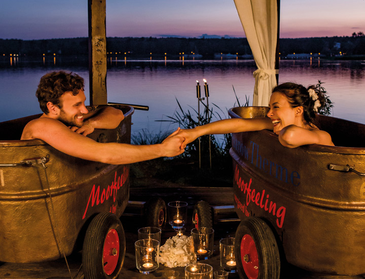 Pavillon am See mit zwei Moorwannen. Im Licht der Sterne und Kerzenschein genießt ein Pärchen ein warmes Moorbad am See. Die Wannen stehen neben einander und das Pärchen geben Sie liebevoll die Hand.