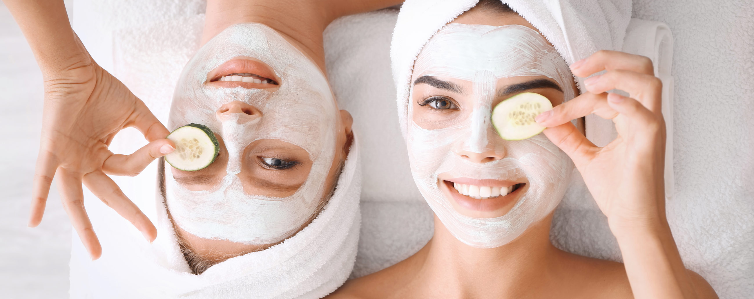 Zwei junge Frauen liegen auf dem Rücken auf einem Bett. Haare sind eingehüllt in einem Handtuch und auf dem Gesicht tragen Sie eine Gesichtsmaske und halten sich jeweils eine frische Gurke vor dem Auge.