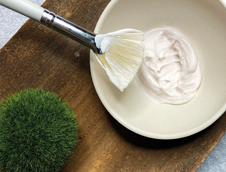 Schale mit cremiger Gesichtsmaske und Pinsel zum Auftragen, dekorativ auf einem Holztablett arrangiert, neben einer kleinen grünen Mooskugel.