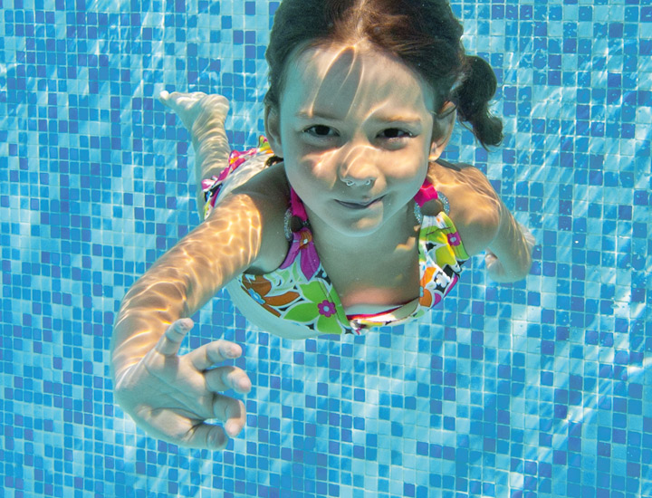 Unterwasseraufnahme eines kleinen Mädchens in einem bunten Badeanzug, dass das Schwimmen lernt. Sie lächelt und winkt in Richtung Kamera.