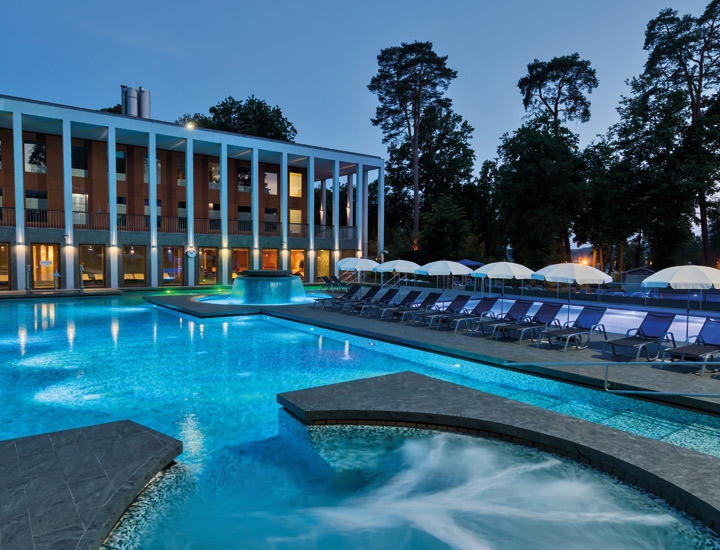 Außenansicht der SaarowTherme bei Abenddämmerung: Beleuchtetes Solebecken mit Massagedüsen, umliegende Liegestühle mit Sonnenschirmen und modernes Gebäude mit Säulen im Hintergrund.