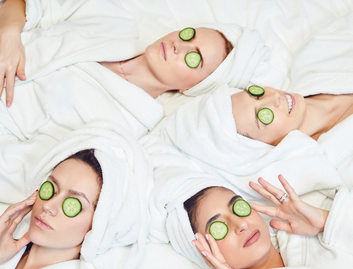 Vier Frauen in weißen Bademänteln und Handtuchturbanen liegen entspannt nebeneinander, mit Gurkenscheiben auf den Augen. Ein klassischer Spa-Moment zur Entspannung und Hautpflege.