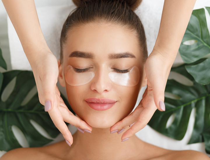 Eine Junge Frau erhält eine Beautybehandlung. Auf dem Rücken liegt Sie auf eine Wellnessliege. Ihr Gesicht mit Augenpads bestückt. Ihre Haare in einem Bun hochgesteckt. Die Hände der Therapeutin streicheln sanft Ihr Gesicht mit pflegenden Produkten. 