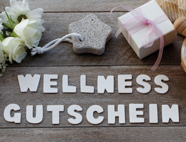 Imagebild mit dem Schriftzug ‚Wellness Gutschein‘ in Weiß im Vordergrund. Im Hintergrund Dekoelemente: weiße Rosen, ein sternförmiger Bimsstein und eine kleine Geschenkbox mit rosa Stoffband. 