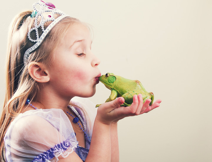Ein Kind in einem Prinzessinnenkostüm mit Krone hält einen grünen Frosch in den Händen und küsst ihn. Die Szene erinnert an das Märchen „Der Froschkönig“ und wirkt verspielt und fantasievoll.