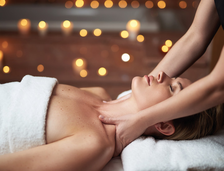 Frau liegt auf dem Rücken auf eine Massageliege eingewickelt in einem Handtuch. Am Kopfende der Massageliege steht eine Masseurin und massiert die Schulter der Frau. 