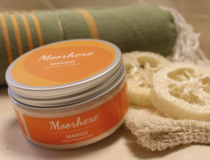 Nahaufnahme eines Peelingsalzes der Marke Moorhexe mit orangem Etikett und der Aufschrift ‚Moorhexe Mango. Im Hintergrund Seifenschwämme und ein grünes Hamamtuch.