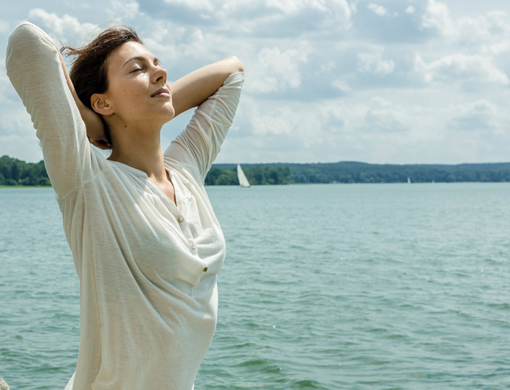 Eine Person steht entspannt am Wasser, hebt die Arme hinter den Kopf und schließt die Augen. Im Hintergrund sind ein See, Bäume und Segelboote unter einem leicht bewölkten Himmel zu sehen – die Szene vermittelt Ruhe und Wohlbefinden.