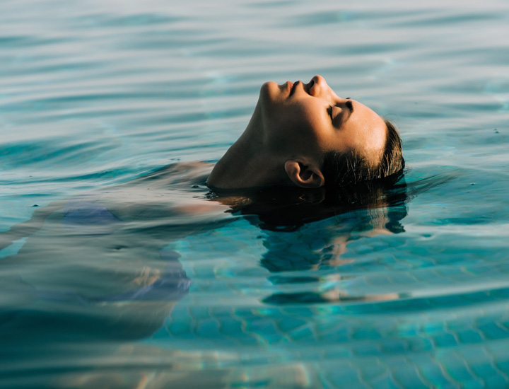 Entspannte Frau im türkisblauen Thermalsolewasser der SaarowTherme, den Kopf nach hinten geneigt, Haare im Wasser treibend, das Gesicht zur Sonne gerichtet und den Moment der Ruhe genießend.