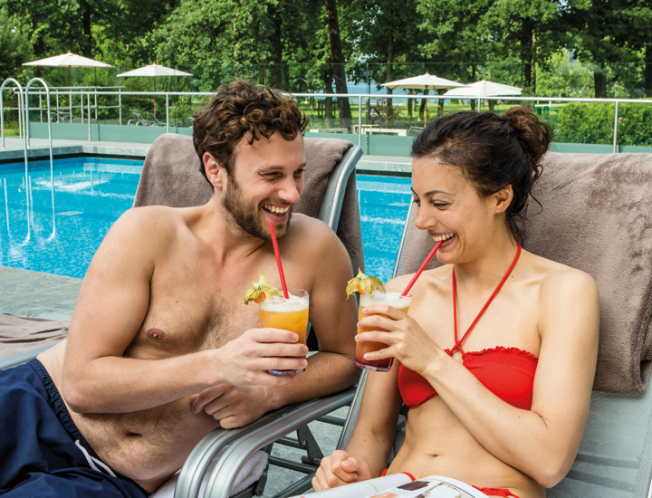 Ein entspanntes Pärchen liegt auf einer Sonnenliege am Outdoorbecken der SaarowTherme und genießt gemeinsam Cocktails. Im Hintergrund erstreckt sich der idyllische, grüne Kurpark von Bad Saarow.