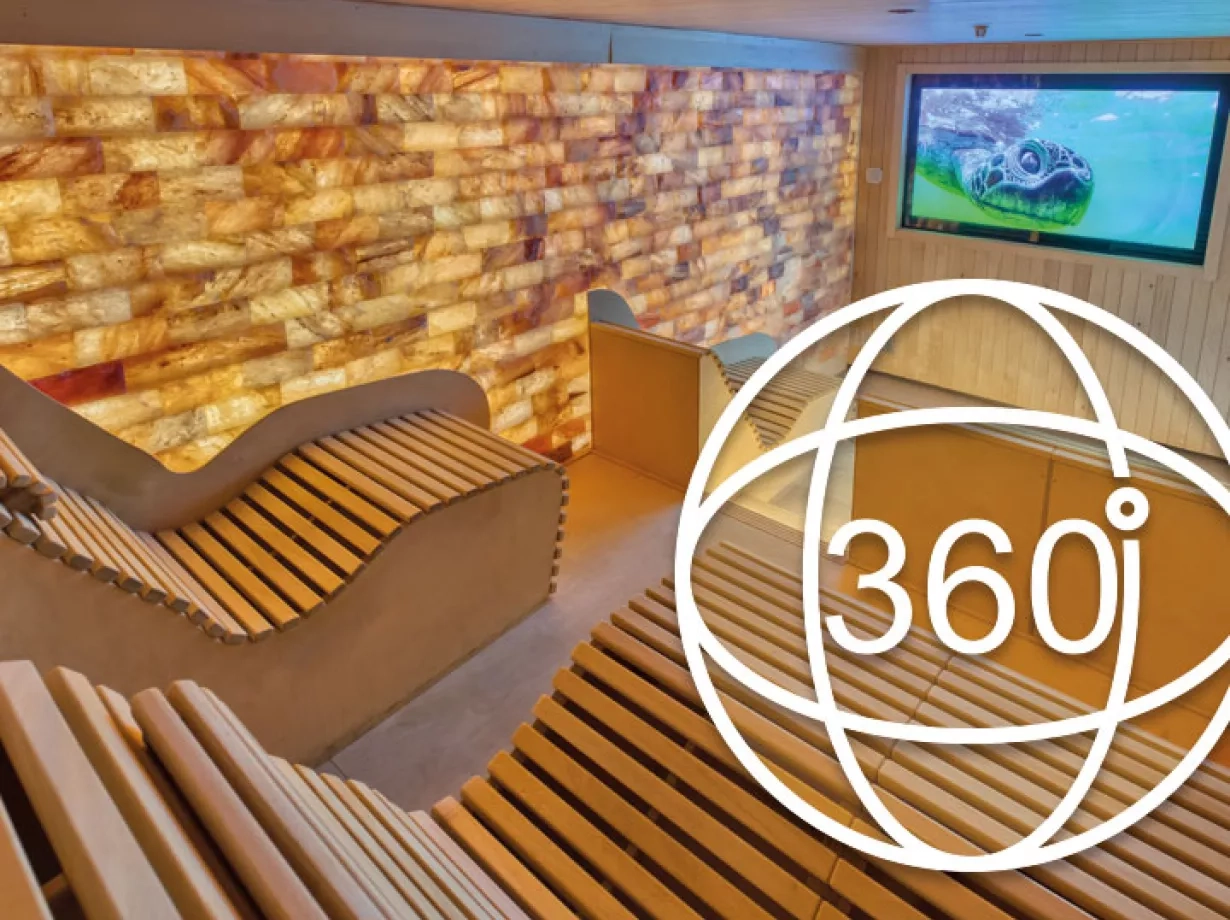 Gemütliche KinoSauna mit beleuchteter Salzsteinwand, ergonomisch geformten Holzliegen und einem Monitor an der Wand, auf dem ein Naturvideo läuft. Im Vordergrund ist das Icon für den 360-Grad Rundgang zu sehen.