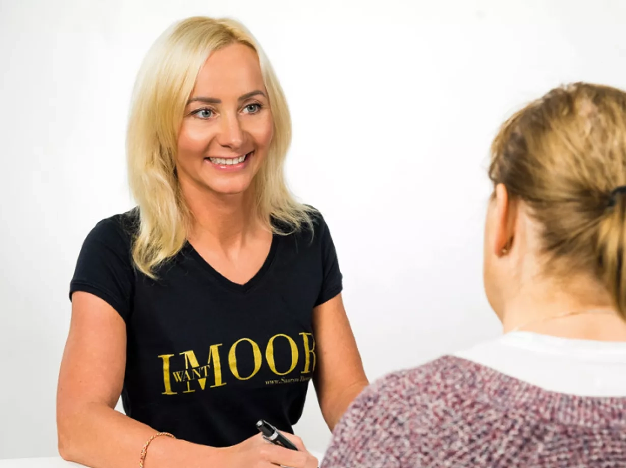 Eine Junge Therapeutin sitz an einem Tisch und hält einen Stift in der Hand. Sie trägt ein schwarzes T-Shirt mit goldenem Schriftzug. Gegenüber der Therapeutin sitzt eine weitere etwas ältere Frau. Sie lächeln sich gegenseitig an.