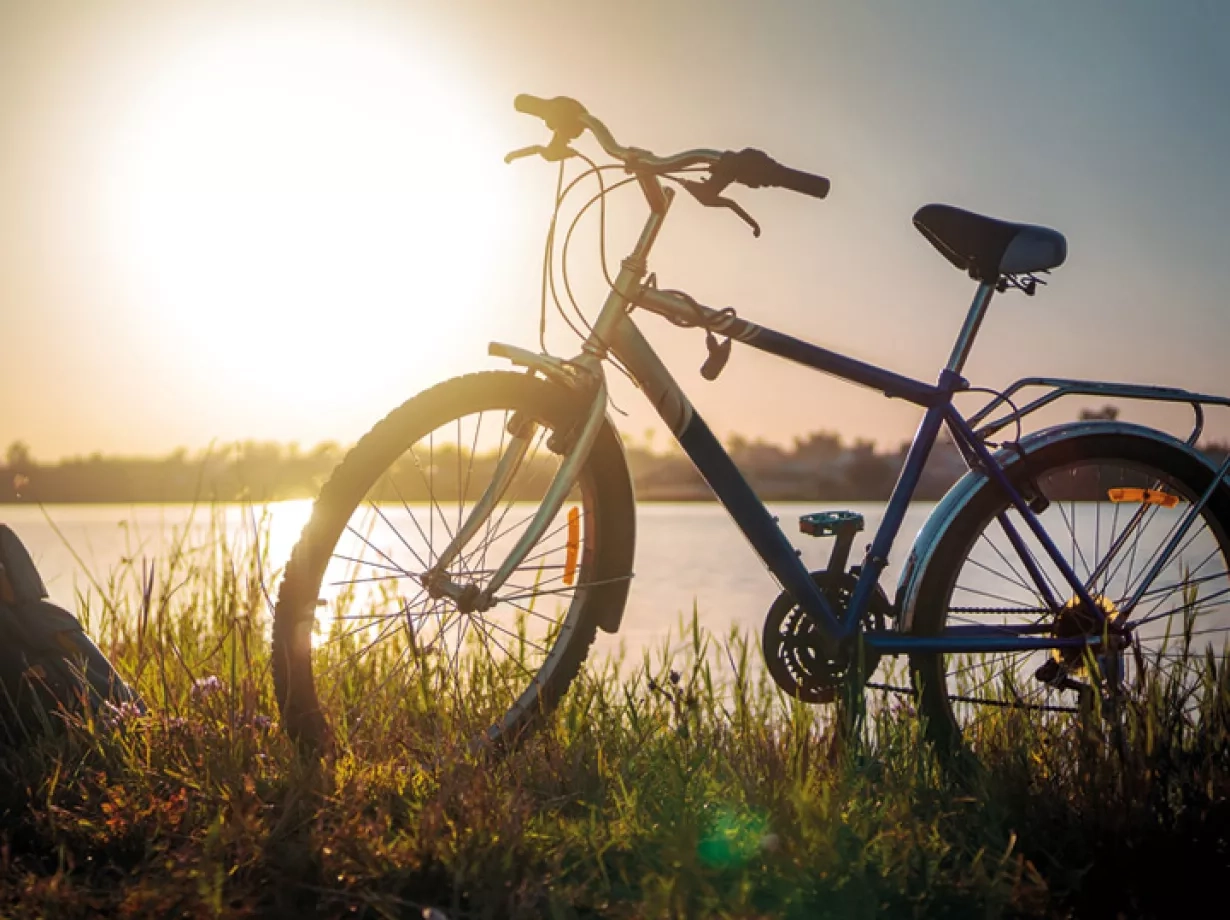 Blaues Sportrad steht am Ufer des Sees. Auf dem grünen Gras neben dem Fahrrad einen blauen Rucksack und im Hintergrund erkennst man einen wunderschönen Sonnenuntergang über dem See.