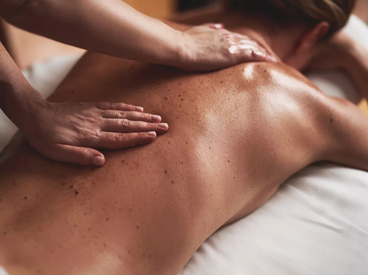 Frau liegt in Bauchlage auf eine Massageliege. Ihre Haare sind leicht zusammengesteckt und der Rücken mit Massageöl eingecremt. Eine Masseurin gibt ihr gerade eine Rückenmassage. 