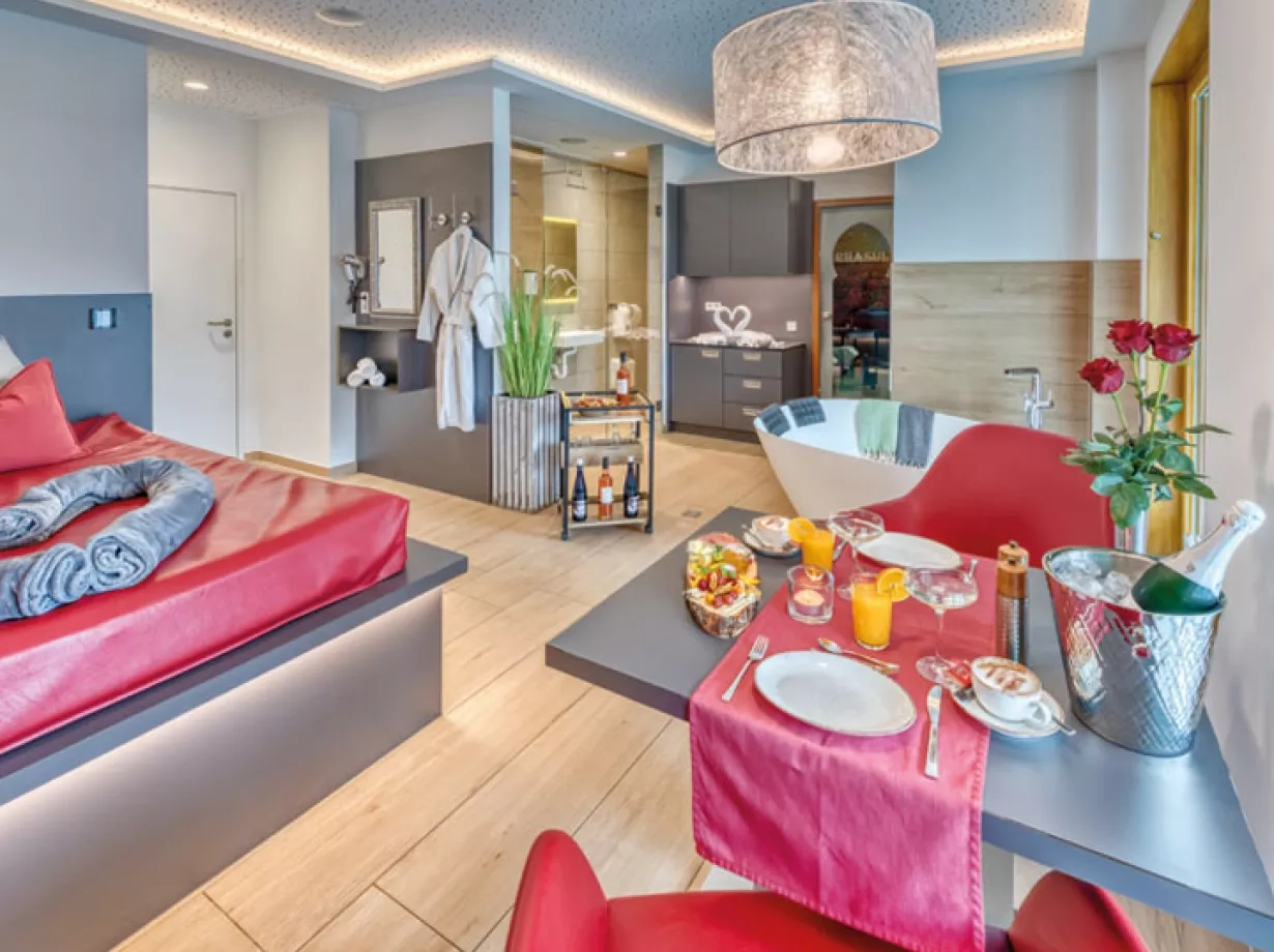 Ausschnitt aus der Private Spa Suite. Im Vordergrund steht ein mit Sektfrühstück bedeckter Tisch. Mitten im Raum steht eine freistehende Duowanne, daneben einen Rollwagen mit Getränken. Im Hintergrund erkennt man das Rhasul und den Sanitärbereich der Suite.