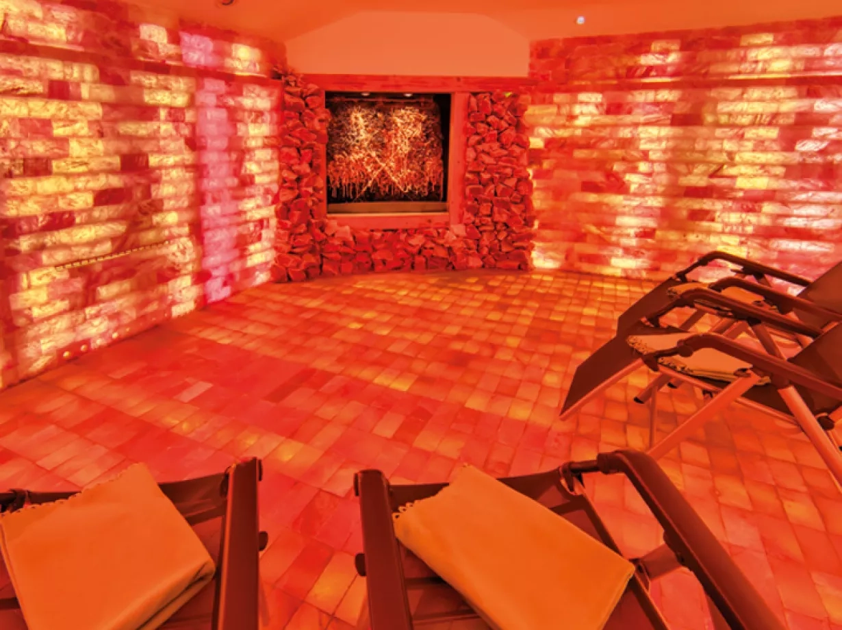 Raum mit warmem orange-rotem Licht, dessen Wände und Boden aus leuchtenden Salzsteinen bestehen. Im Hintergrund ist ein beleuchtetes Wandbild zu sehen, im Vordergrund stehen mehrere Liegestühle mit Handtüchern – der Raum wirkt ruhig und entspannend, typisch für eine Salzgrotte oder einen Wellnessbereich.