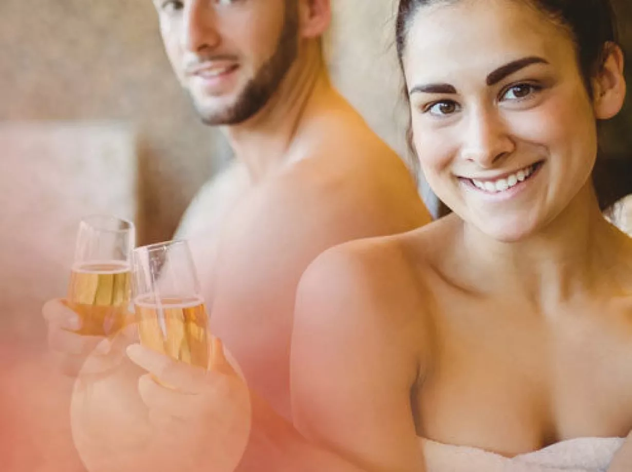 Ein Mann und eine Frau in Handtüchern sitzen entspannt in einer Wellness-Umgebung und halten Gläser mit Sekt oder Saft in der Hand. Beide lächeln in die Kamera.