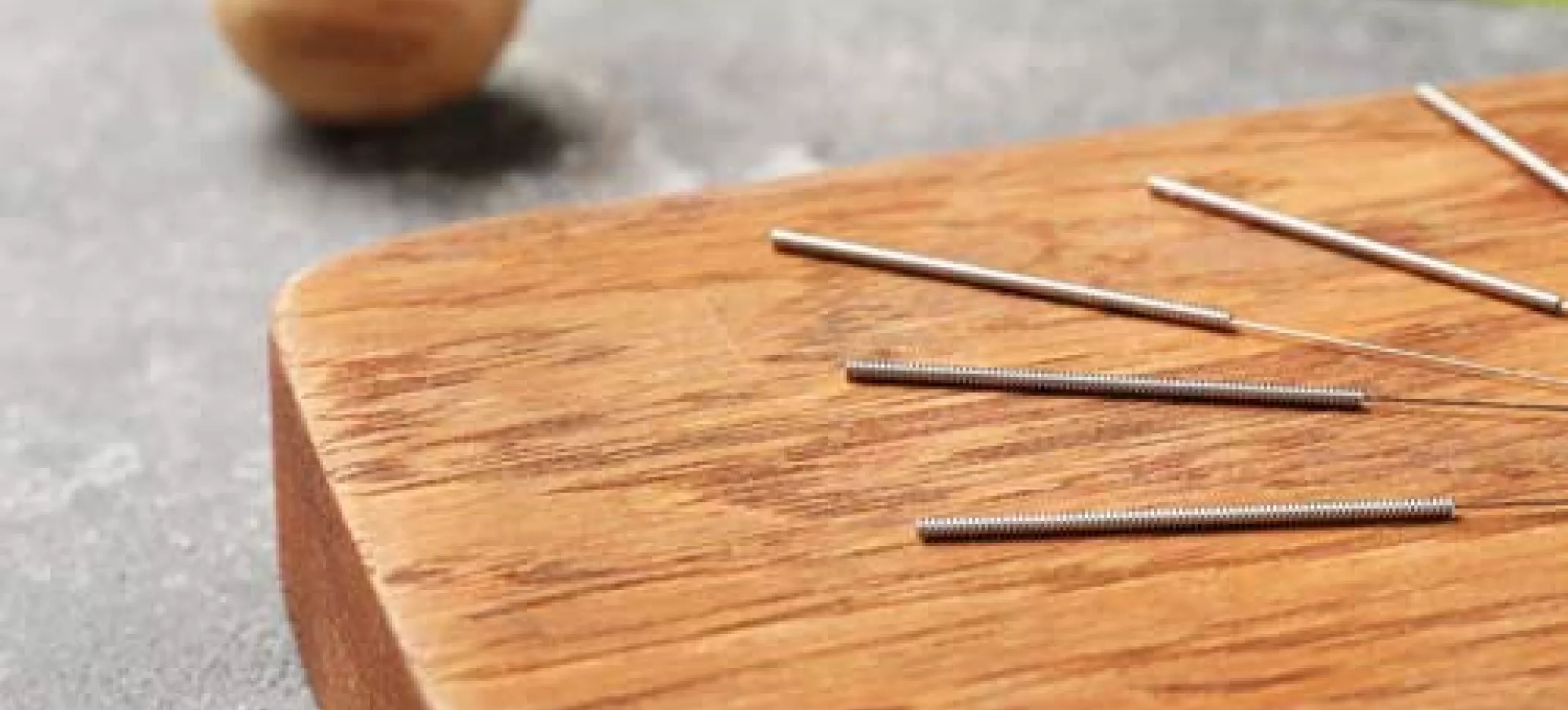 Ausschnitt auf dem eine helle Holzfläche zu sehen ist, die auf einem grauen Untergrund platziert ist und auf dieser Holzfläche befinden sich hauchdünne Akupunkturnadeln. Dekorativ befinden sich Grünpflanzen im Hintergrund. 