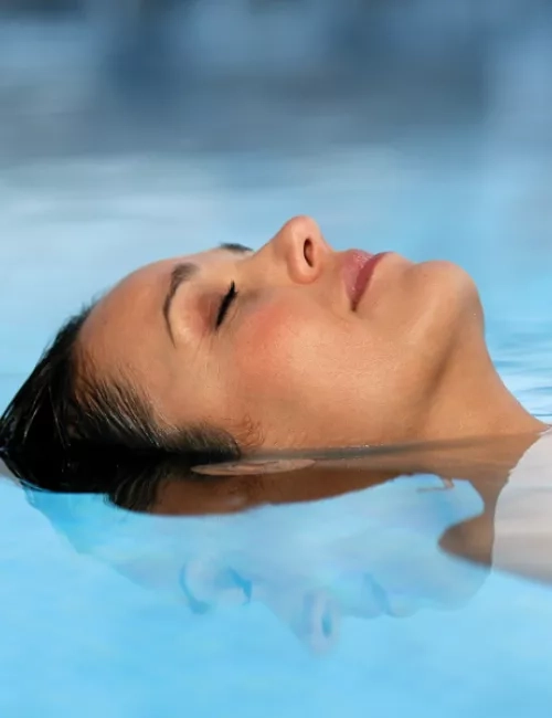 Auf dem Wasser schwebende Frau mit dunklen Haaren, der Kopf über Wasser mit dem Gesicht nach oben und geschlossenen Augen. Nahaufnahme, sodass der Kopf und nur ein Teil des Oberkörpers zu sehen ist. Das Wasser indem Sie liegt ist hell blau.
