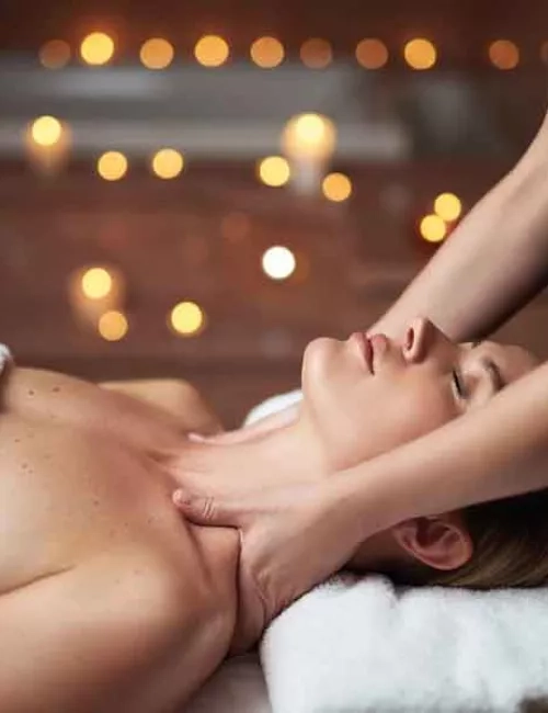 Ein auf den Rückenliegende Frau, wird im Nacken-Schulterbereich massiert. Ihr Kopf liegt auf einem weißen, flauschigen Handtuch und im Hintergrund sind verschwommene, angezündete Kerzenlichter zu erkennen. Die Augen der behandelten Frau sind geschlossen und Ihr Gesicht wirkt sehr entspannt.