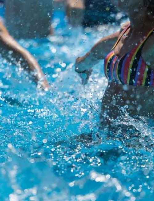 Kleiner Ausschnitt eines Bildes, mit Türkisfarbendes Wasser in der eine Frau mit einem schwarz,blau, rot,gelb längst gestreiften Bikinioberteil und weit geöffneten Armen hüpft und somit das Wasser in Bewegung bringt. 