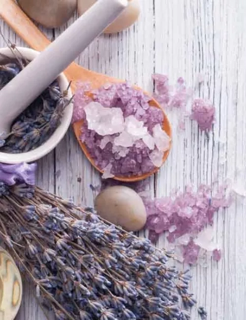 Produktbild von einem Lila Badezusatz in einer groben Salzform auf einem Holzlöffel platziert. Dekorativ befinden sich darum Lavendelbüsche und etwas loses Badesalz.