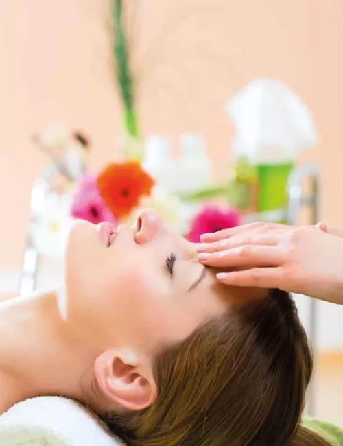 Eine Frau mit dunkelbraunen Haaren und einem Zopf liegt auf eine Massageliege, im Nacken wird der Kopf mit einer weißen Nackenrollen gestützt. Ihre Augen sind geschlossen und auf Ihrer Stirn liegen zart zwei Hände auf und deuten eine Massage an. Im Hintergrund befinden sich dekorative Elemente in Form von Grünpflanzen. 