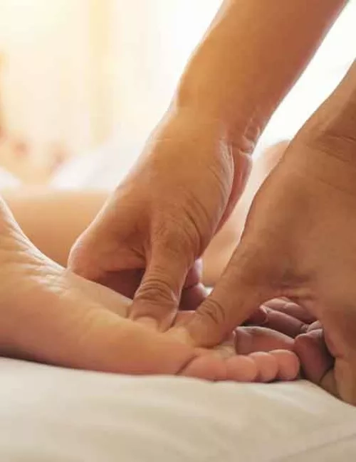 Füße die auf einem weißen, weichen Untergrund liegen. Die Fußsohlen zeigen nach oben und zwei Hände berühren ein Fuß. Die Daumen drücken in die Fußsohle des linken Fußes. Helles Tageslicht im Hintergrund. 