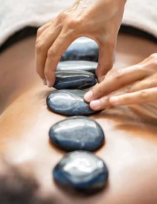 Eine Person liegt auf dem Bauch und bekommt von zwei Händen graue Hot Stone Steine auf die Wirbelsäule gelegt. Nahaufnahme.