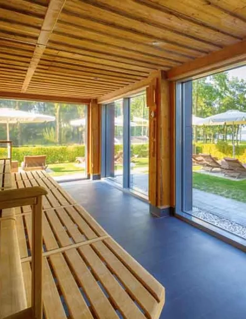 Abgebildet ist die KeloSauna der PanoramaSauna mit Blick auf den grünen Kurpark in Bad Saarow, mit Blick auf den blauen Scharmützelsee. Die Saunadecke und die Saunabank, sind mit groben Holzstrukturen versehen. Der Fußboden besteht aus dunkel grauen Fließen. Eine riesige Glasfront lässt es zu, dass der Raum Sonnendurchflutet wird und man sieht die braunen Außenliegen mit weißen aufgespannten Sonnenschirmen. Ein dunkel grauer Saunaaufgussofen ist am Rande des Bildes zu erkennen.