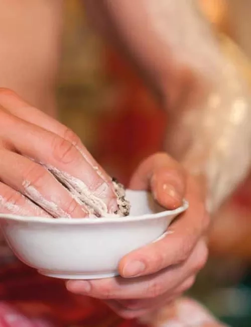 Eine sitzende Person mit einem roten Hamamtuch und im Vordergrund wird eine weiße Schale mit einem Naturerdenpeeling gehalten. Die rechte Hand greift in die Schale und auf dem linken Arm befindet sich bereits das Naturerdenpeeling.