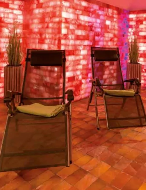 Abgebildet ist die Salzoase mit einem Fußboden und Wänden aus Salzsteinen, welche in verschiedenen, warmen Rosafarbenen Tönen beleuchtet werden. Im Vordergrund stehen zwei Liegen mit grünen Kuscheldecken. Dekorativ im Hintergrund befinden sich zwei Grünpflanzen stehend in einem länglichen Übertopf. 