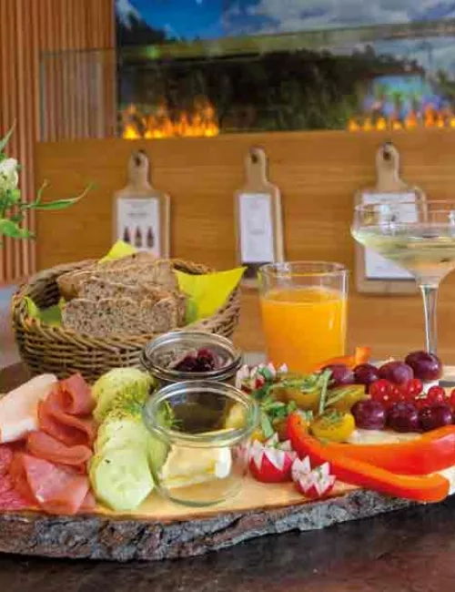 Ein rustikale Holzplatte mit verschiedenen Speisen, wie Gurke, rote Paprika, Butter und Marmelade im kleinen Einwegglas und Salami, dekorativ angerichtet. Ein Brotkorb mit Brotscheiben und ein Glas Orangensaft und ein Glas Sekt. Im Hintergrund sind künstliche Flammen zu sehen. 