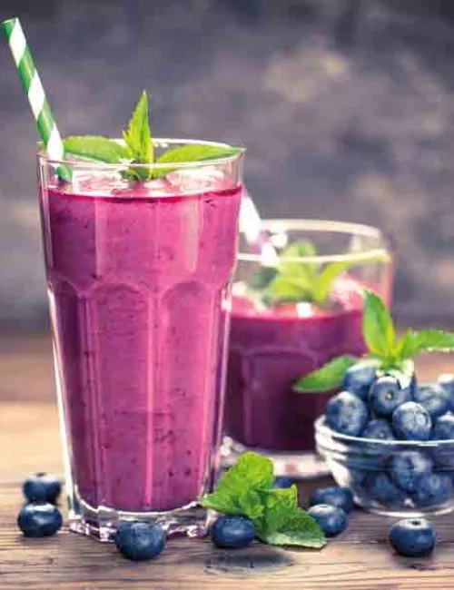 Im Vordergrund befindet sich ein großes Glas mit einem Lilafarbenen Smoothie. Im Hintergrund ein kleineres Glas ebenfalls mit einem lilafarbenen Smoothie Inhalt. Dekorativ ist eine kleine Glasschlüssel mit Heidelbeeren und grünen Minzblättern platziert. Vereinzelt wurden einige Heidelbeeren und Minzblättern vor den Gläsern platziert.