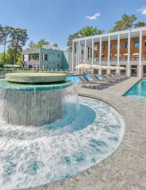 Außenbereich des Thermalsolebeckens  SaarowTherme am Tag mit plätschernden Strömungskanal, graue Liegen mit weißen aufgespannten Sonnenschirmen. Im Hintergrund ein Teil des Hauptgebäudes mit hellbrauner Gebäudefront und weißen Säulen im Vordergrund. Blauer Himmel mit weißen Schleierwolken. 