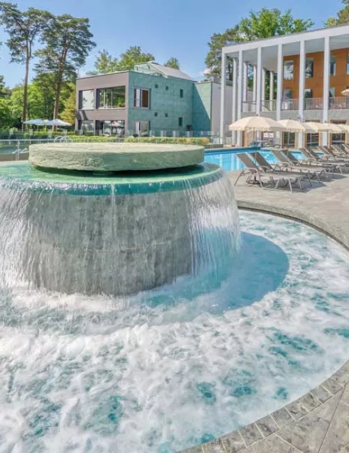 Außenbereich des Thermalsolebeckens  SaarowTherme am Tag mit plätschernden Strömungskanal, graue Liegen mit weißen aufgespannten Sonnenschirmen. Im Hintergrund ein Teil des Hauptgebäudes mit hellbrauner Gebäudefront und weißen Säulen im Vordergrund. Blauer Himmel mit weißen Schleierwolken. 